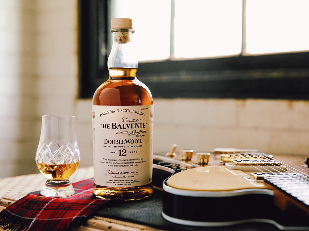 A glass and bottle of Balvenie Double Wood Single Malt Scotch Whisky set on a table beside an electric guitar