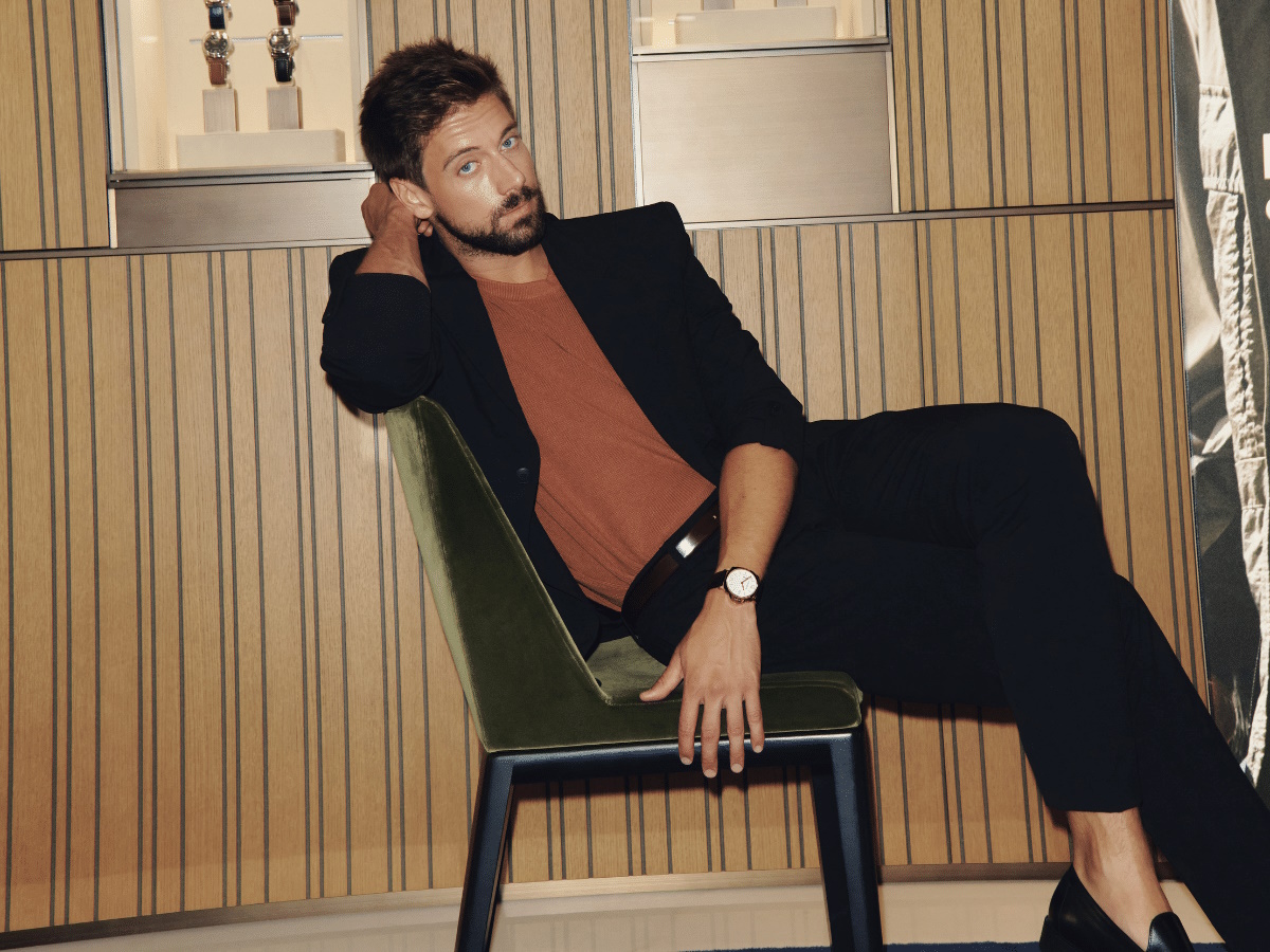 Male model in a brown shirt, black jacket and pants, languidly sitting on a chair