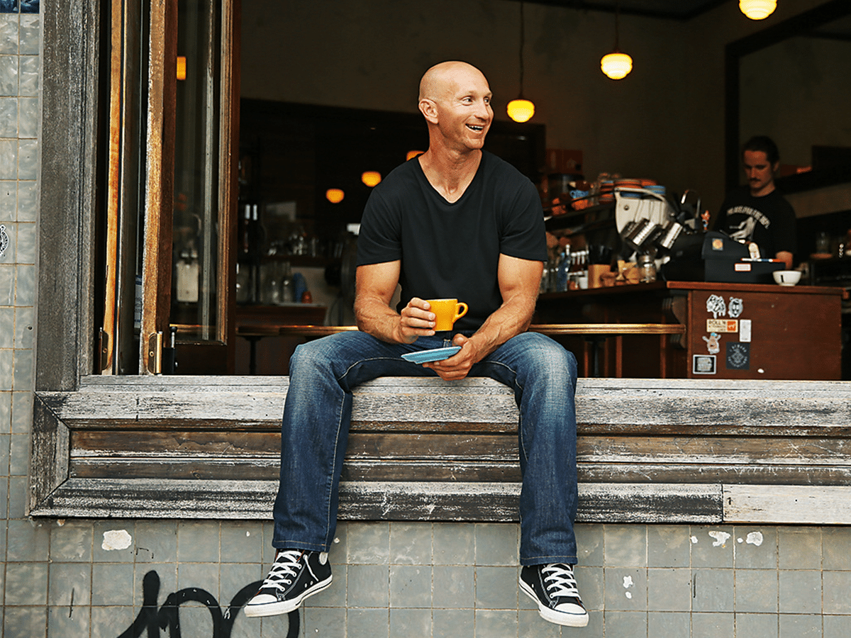 Man in a black t-shirt, jeans, and converse sneakers holding a teacup while sitting on a window sill